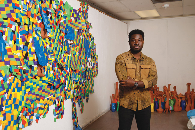 artist standing in studio next to multi-color wall aculpture
